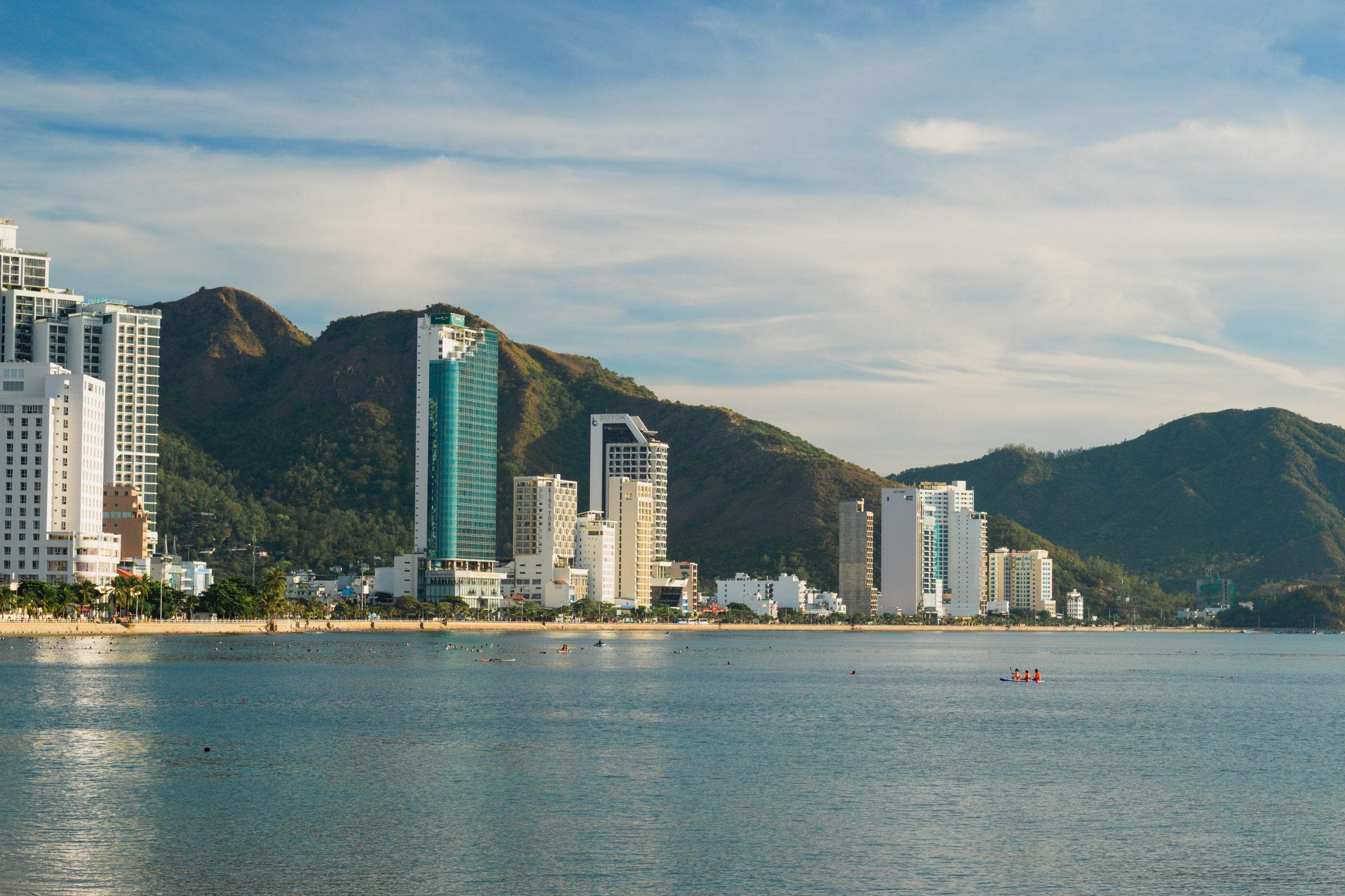 City skyline with skyscrapers and mountains reflecting on calm coastal waters.