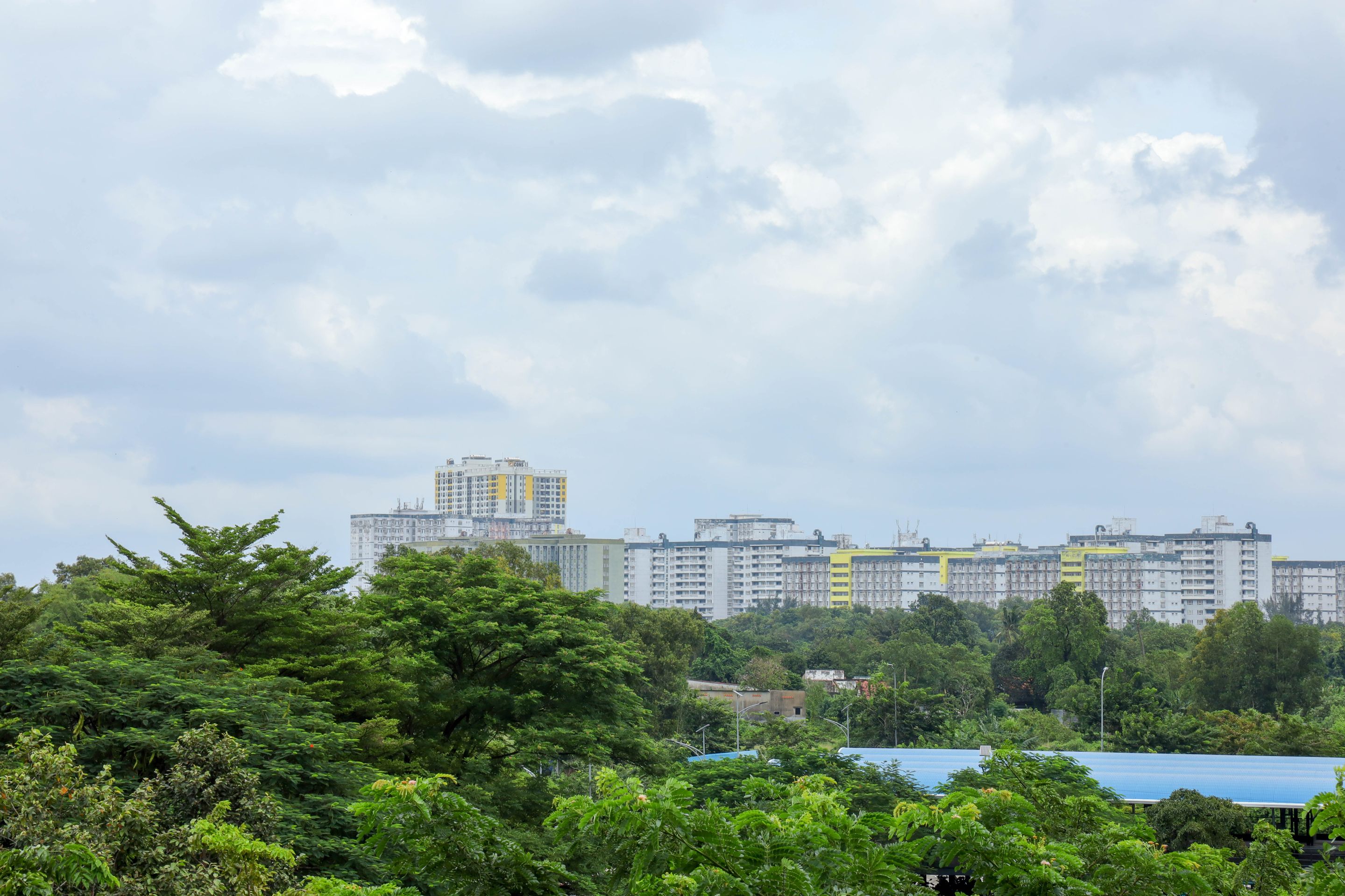 Urban cityscape featuring high-rise residential buildings with lush greenery in the foreground under a cloudy sky.