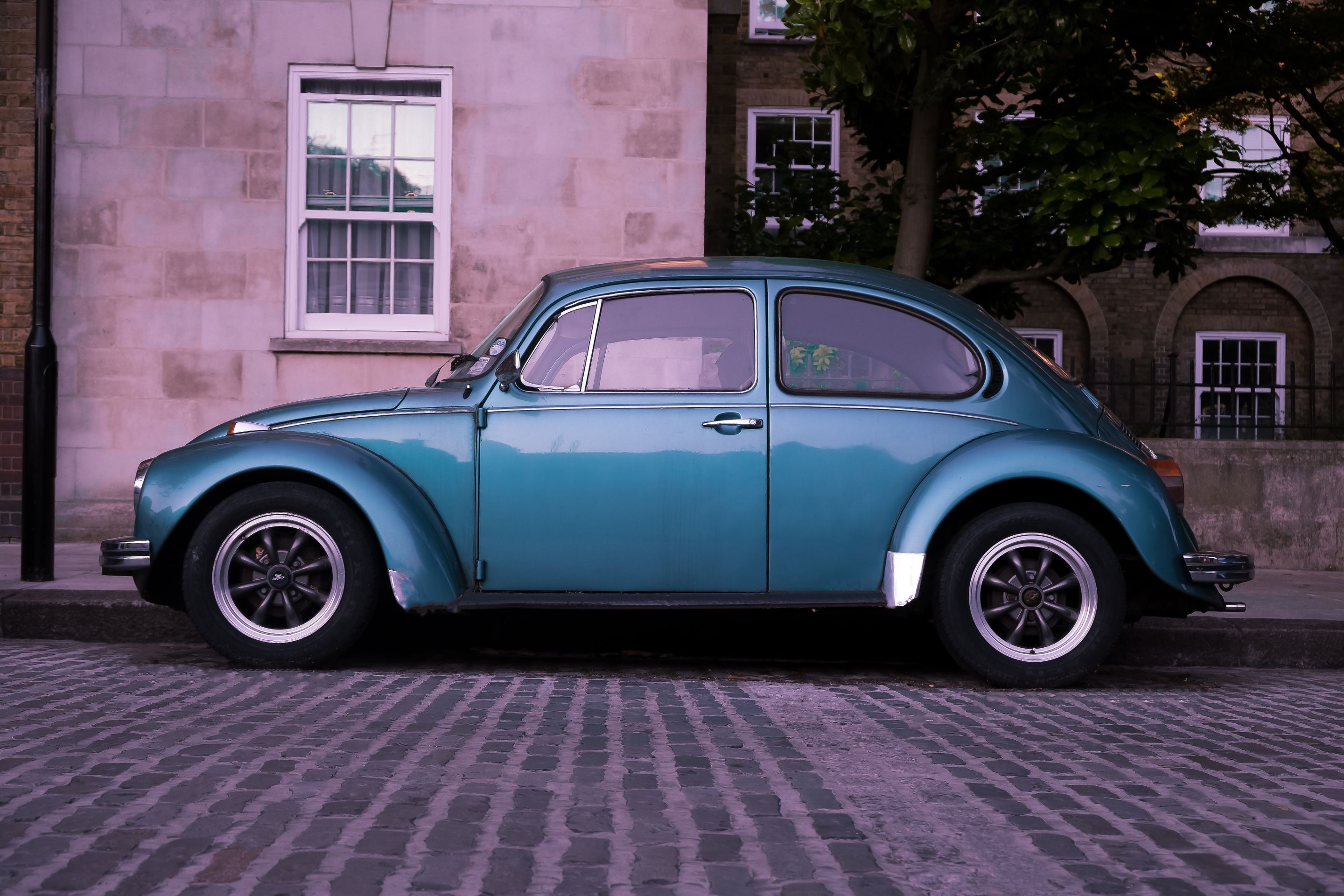 Classic turquoise Volkswagen Beetle parked on a cobblestone street with a brick building backdrop.